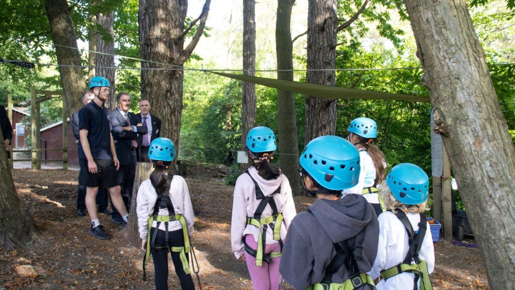 Young people looking and engaging with outdoor learning apprentice wearing helmets and harnesses in the forest under trees ready to do tree top trail